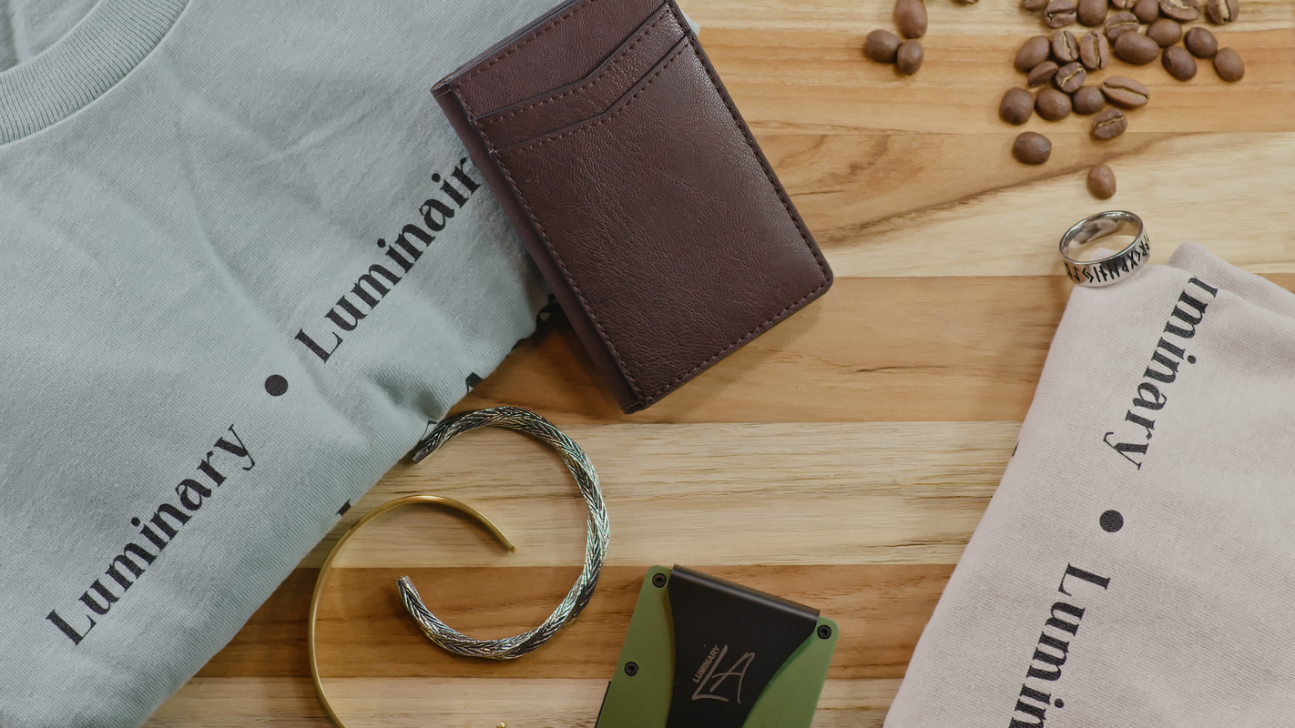 Flat lay shot of LuminaryLA products against a wood panel background with coffee beans. Products include a light blue hoodie, tan shirt, brown leather wallet, card holder, gray and silver bracelets, and silver ring.